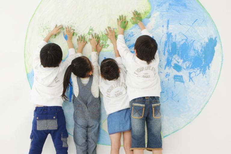 Four children stand facing a wall, pressing their painted hands onto a large drawing of the Earth.