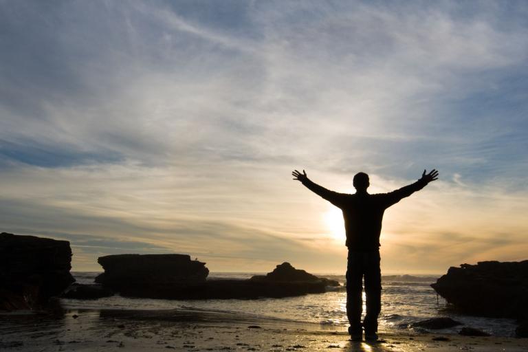 A person stands on a rocky beach with arms outstretched, facing the ocean at sunset under a partly cloudy sky.