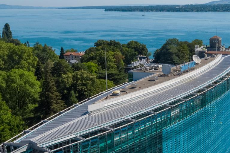 A modern curved glass building with solar panels on the roof, surrounded by trees, near a large lake with distant hills in the background.
