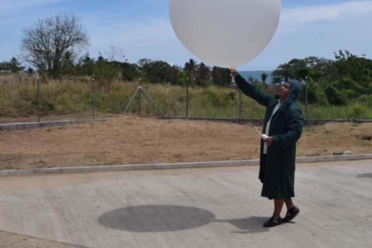 A person in a coat and headscarf stands on a paved area holding a large white weather balloon outdoors under a blue sky.