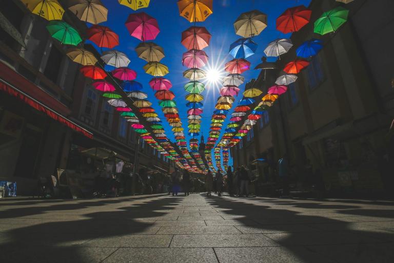 Colorful umbrellas are suspended above a pedestrian street, casting shadows on the ground; sunlight shines through the umbrellas and buildings line both sides of the street.