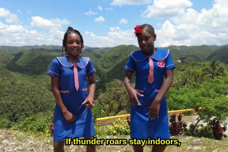 Two girls in blue school uniforms stand outdoors on a hilltop with green hills in the background. Subtitle reads, "If thunder roars stay indoors," in yellow text.