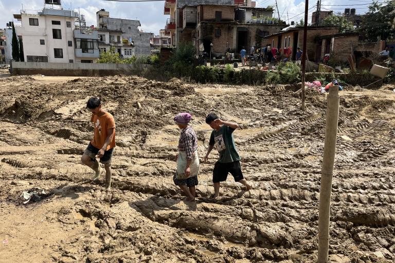 Three people walk through deep mud with building debris and tire tracks in an urban area, with residential houses and onlookers in the background.