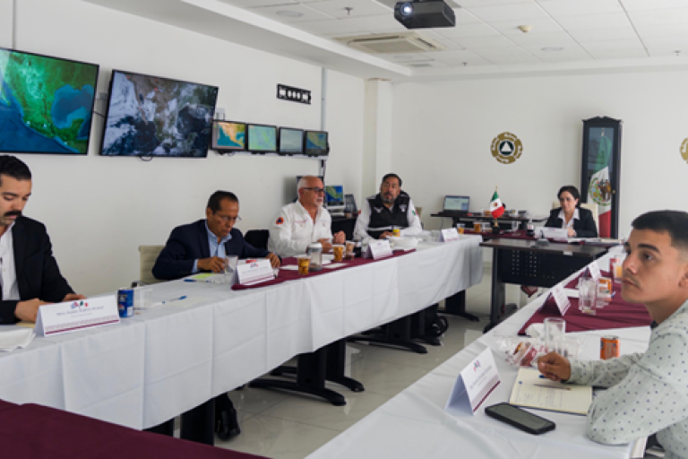 A group of people sit around a conference table in a meeting room with multiple monitors displaying maps on the wall.