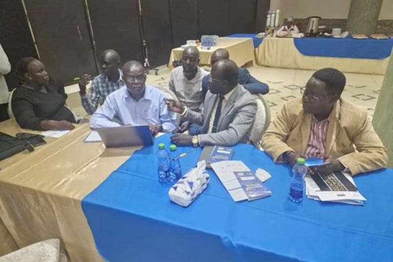 A group of people sit around a table with laptops, notebooks, and bottled water, engaged in discussion at an indoor event or meeting.