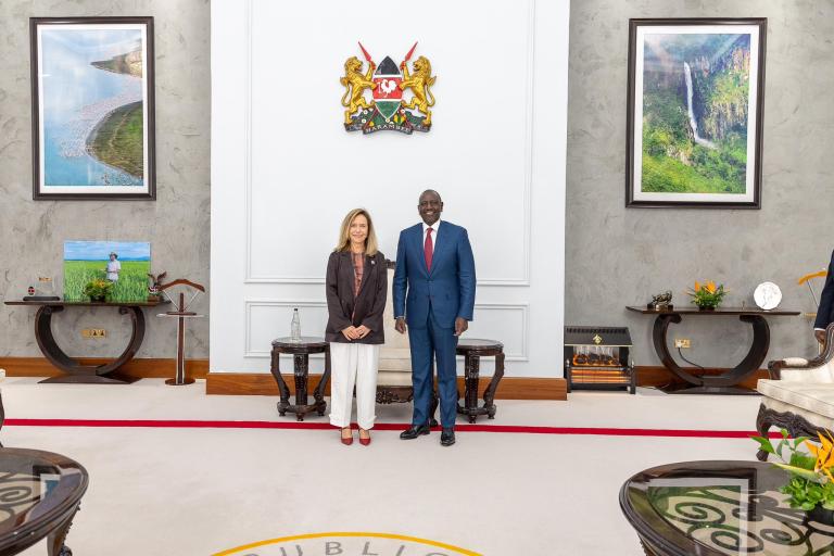 Two people stand side by side in a formal room with framed photographs, decorative furniture, and a national emblem on the wall behind them.