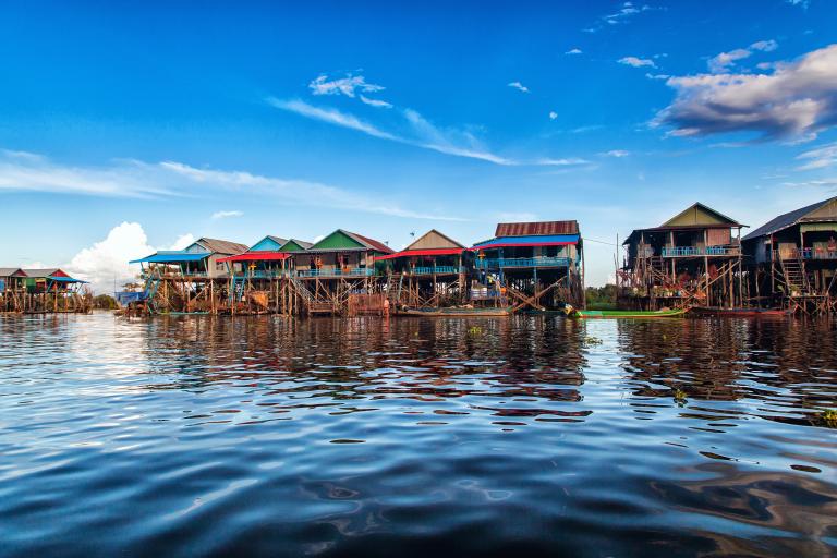 Colorful wooden houses on stilts line the edge of a calm body of water under a blue sky with scattered clouds.