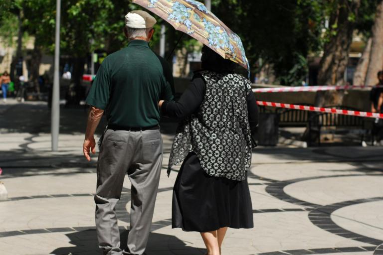 An older couple walks down a sunny, tree-lined sidewalk. The woman holds a large umbrella, and they are walking arm in arm away from the camera.
