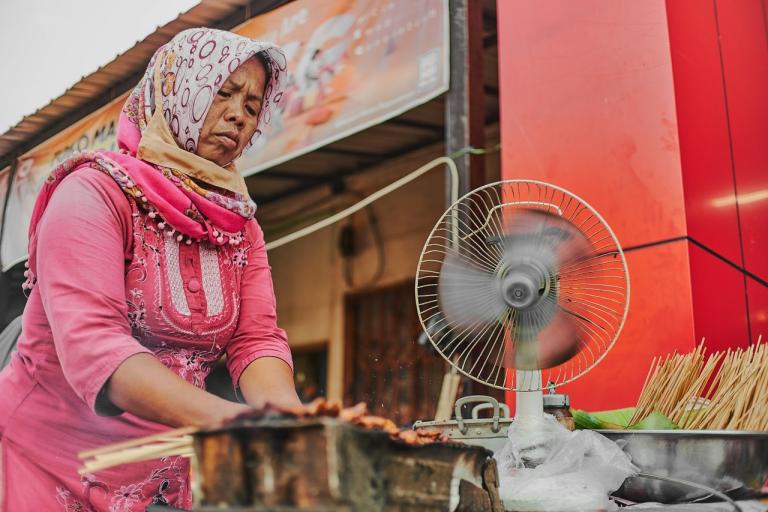 A woman in a pink dress and headscarf grills skewered food beside a spinning fan at an outdoor food stall.
