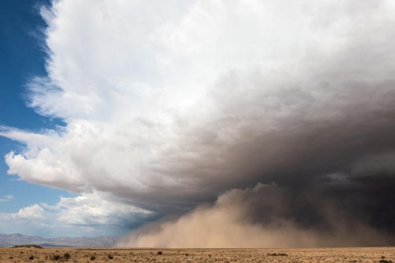 A large dust storm approaches across a dry, flat desert landscape under a partly cloudy sky.