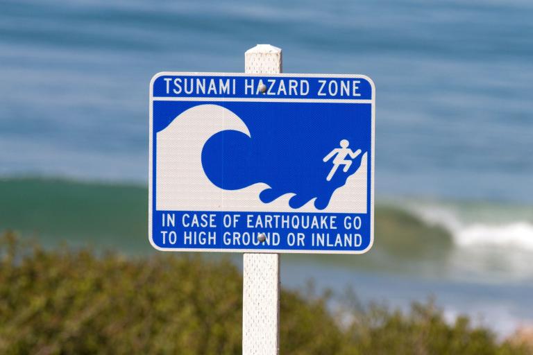 Blue and white sign reads "Tsunami Hazard Zone" and instructs to go to high ground or inland in case of earthquake, with wave and person graphic. Ocean is visible in the background.