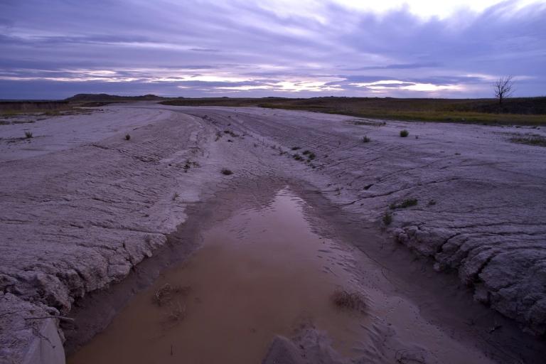 A muddy, shallow stream flows through a dry, cracked landscape under a cloudy sky at dusk, with minimal vegetation and a lone tree in the distance.