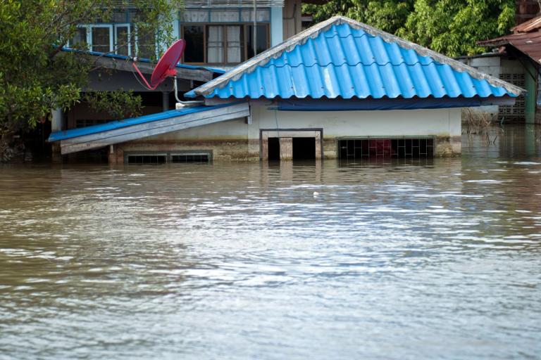 A house with a blue roof is partially submerged in floodwater, with only the roof and upper windows visible above the waterline.