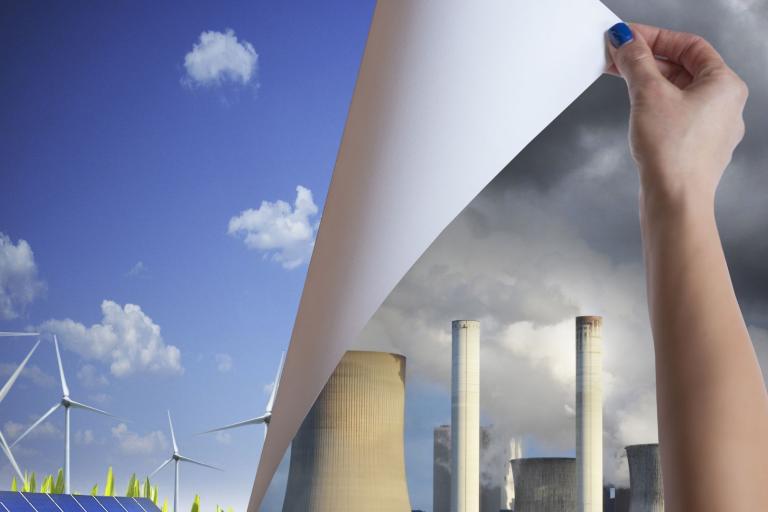 A hand peels back an image of smokestacks and pollution to reveal wind turbines and solar panels under a clear blue sky with clouds.