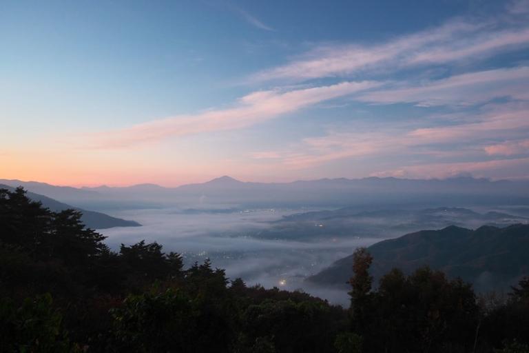 A landscape view of a valley covered in morning mist, with distant mountains under a colorful sunrise sky and scattered lights visible below.