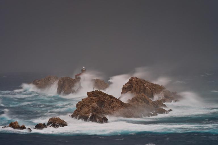 Waves crash against rocky outcrops in a stormy sea, with a lighthouse visible on one of the rocks under a dark, cloudy sky.