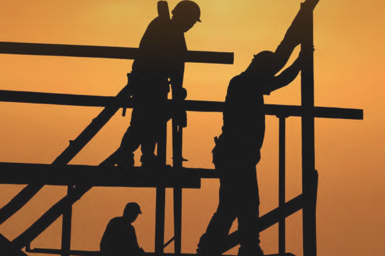 Three construction workers are silhouetted against an orange sunset sky as they work on a building framework.