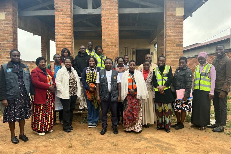 A group of adults stand together outside a brick building, some wearing reflective vests and others in patterned clothing, posing for a group photo.
