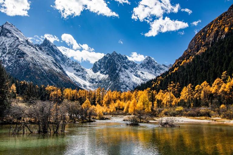 A clear lake reflects yellow autumn trees and snow-capped mountains under a bright blue sky with scattered clouds.