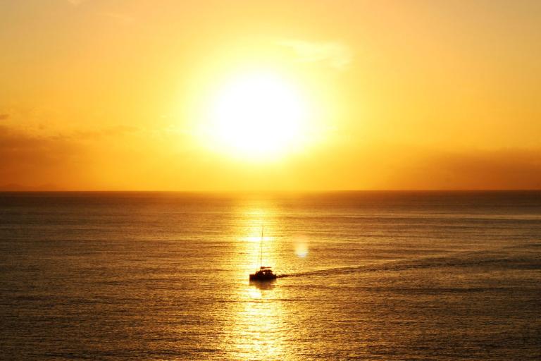 A small boat sails on calm water at sunset, with the sun low on the horizon and golden light reflecting on the sea.