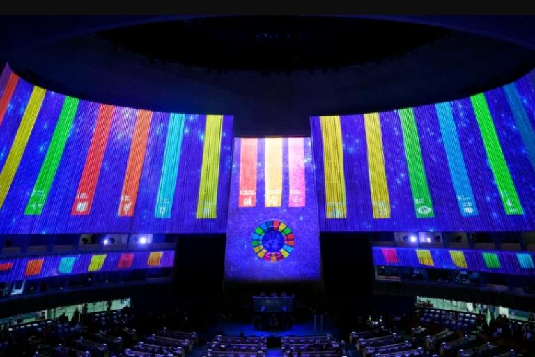 The interior of a large auditorium displays multicolored vertical light panels and a circular emblem on the wall above seated attendees.