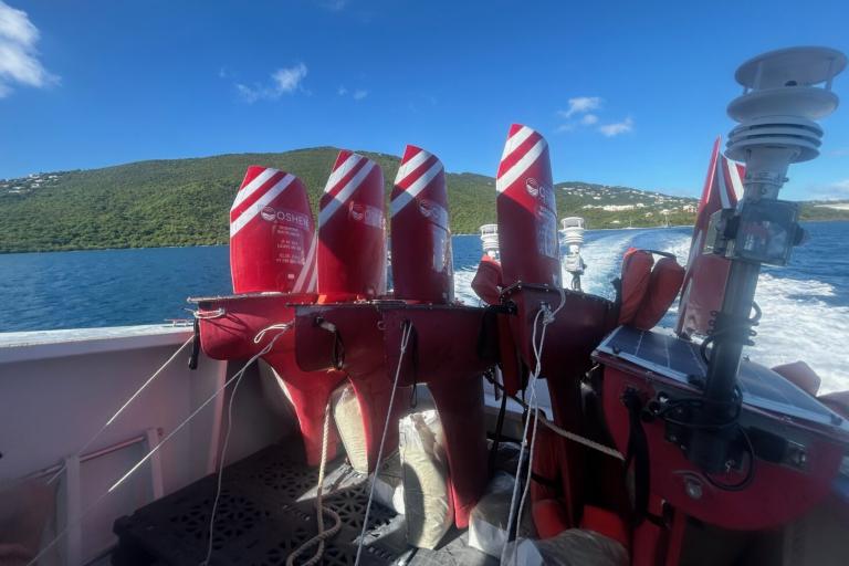 Several red and white Wave Glider marine drones are secured on the back of a boat, with the ocean and a green, hilly coastline visible in the background.