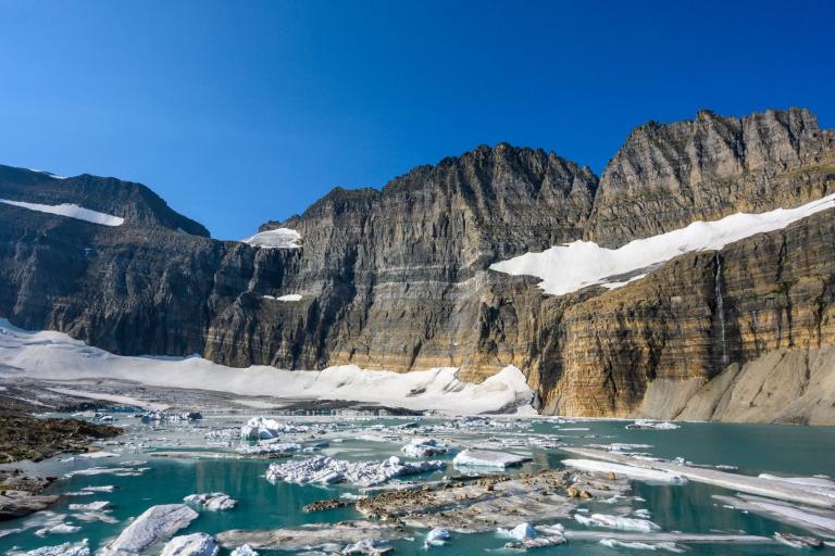 Rocky cliffs rise above a partially frozen turquoise lake with floating ice and patches of snow under a clear blue sky.