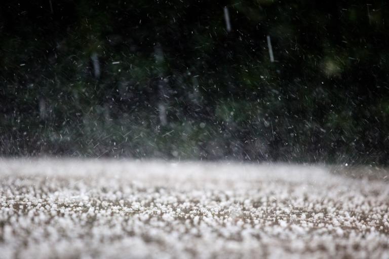 Close-up of hailstones falling and accumulating on the ground, with blurred greenery in the background.