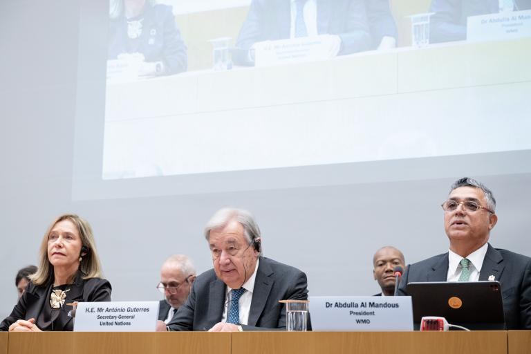 Three officials sit at a conference table with microphones and nameplates; a projected image of the panel is shown above them.