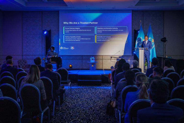 A speaker presents information on a screen titled "Why We Are a Trusted Partner" to an audience seated in a conference room with blue lighting.