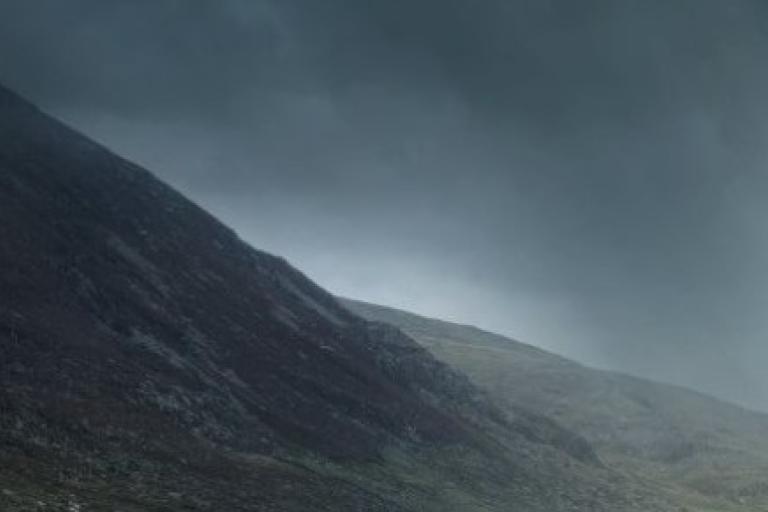 Dark, overcast sky above a sloping, rocky hillside with patches of grass and sparse vegetation.