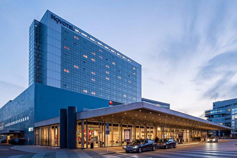 Large modern hotel building with a "Novotel" sign, glass windows, and a covered entrance where cars are parked; city buildings are visible in the background at dusk.