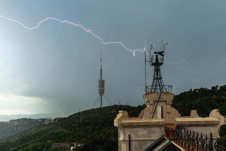 A bolt of lightning strikes near a tall communications tower and an observation building on a hill, with dark clouds overhead and green landscape below.