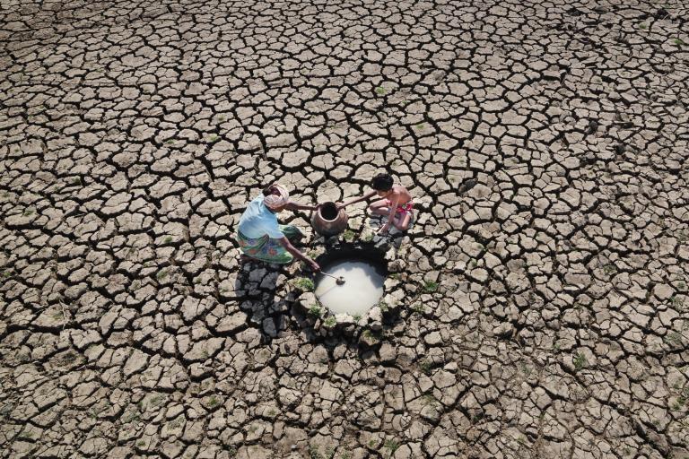 Three people collect water from a small well in the middle of a cracked, dry landscape, indicating drought conditions.