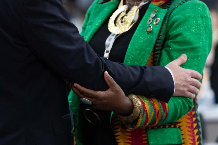 Two people in formal attire embrace; one wears a vibrant green jacket with patterned sleeves and large jewelry, while the other's suit is dark and plain.
