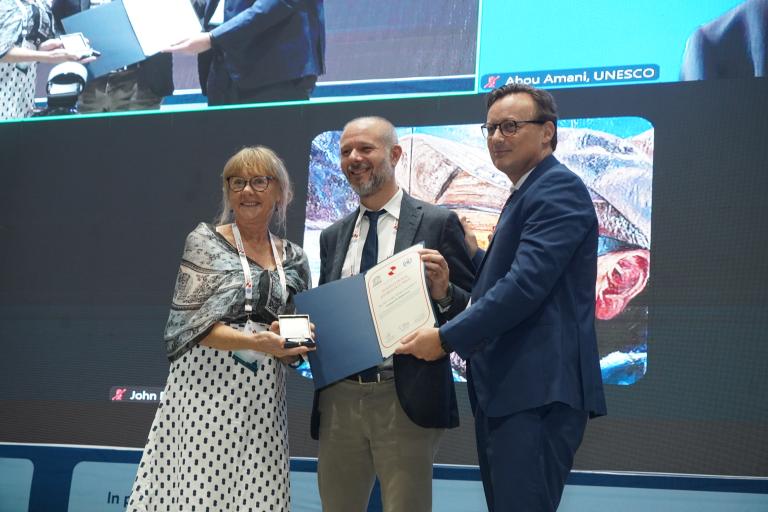 Three people stand on stage holding certificates and an award, with a large screen displaying their image in the background.