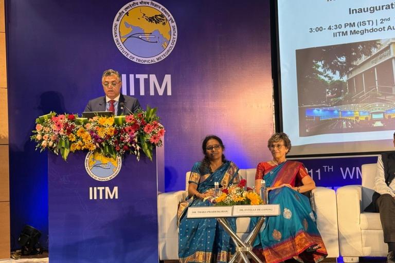 A man speaks at a podium decorated with flowers, with two seated women on stage beside him at an IITM scientific conference event. A presentation slide is visible in the background.