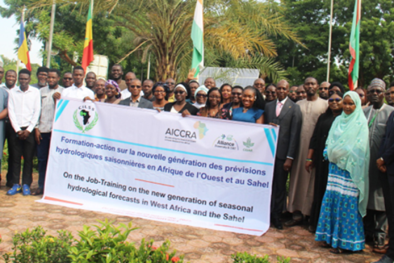 A large group of people pose outdoors, holding a banner about hydrological forecast training in West Africa and the Sahel, with flags and trees in the background.