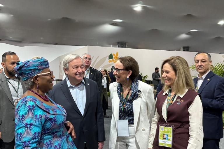 A group of five people, including two women in business attire and one in a blue patterned outfit, stand together indoors, conversing and smiling at an event.