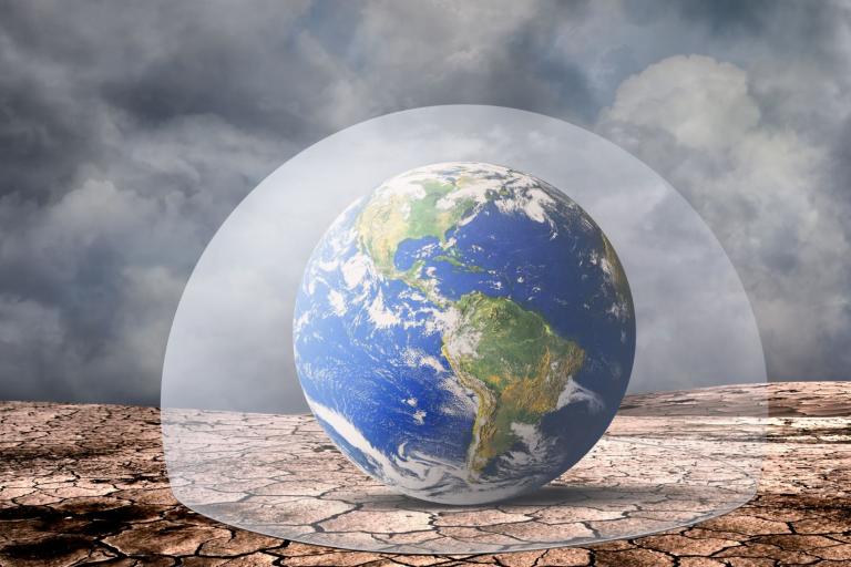 A glass dome encloses a globe showing the Americas, set on cracked, dry ground under a cloudy sky.