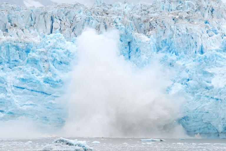 Large chunk of glacier ice collapsing into the water, creating a splash and mist at the base, with blue and white ice formations in the background.