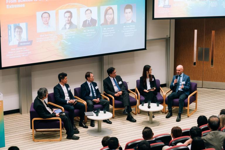 A panel of six people in business attire sit on stage discussing during a conference, with an audience watching and a presentation screen displayed above them.