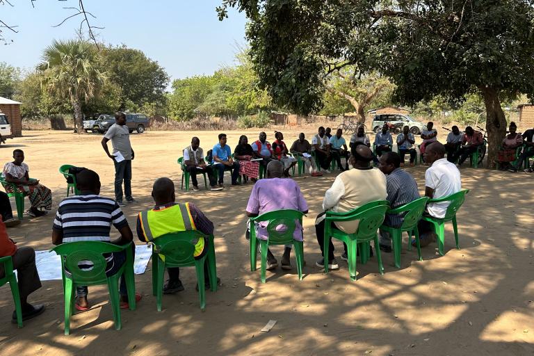 A group of people sit in a large circle on green plastic chairs outdoors, while one person stands and speaks; trees and vehicles are visible in the background.