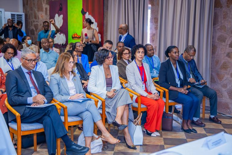 A group of people in business attire are seated in rows at an indoor conference or meeting, listening attentively to a presentation.
