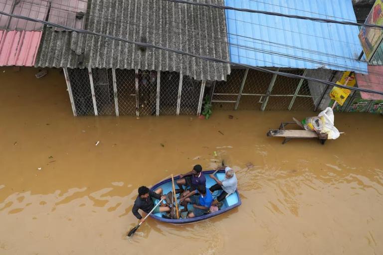 Three people row a small boat through floodwaters in front of submerged buildings, while a man sits on a table with bags nearby.