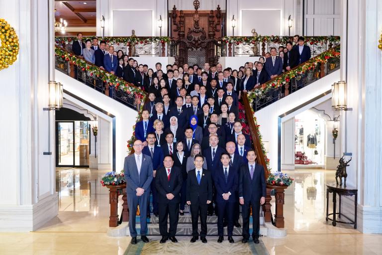A large group of formally dressed people pose for a group photo on a grand staircase decorated with festive garlands in an ornate building lobby.