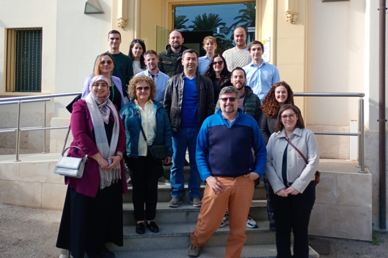 A group of sixteen adults stands on steps outside a building entrance, posing for a photo on a sunny day.