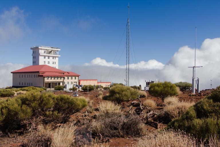 A research observatory building with a red roof, surrounded by dry shrubs and antennas, under a blue sky with scattered clouds.