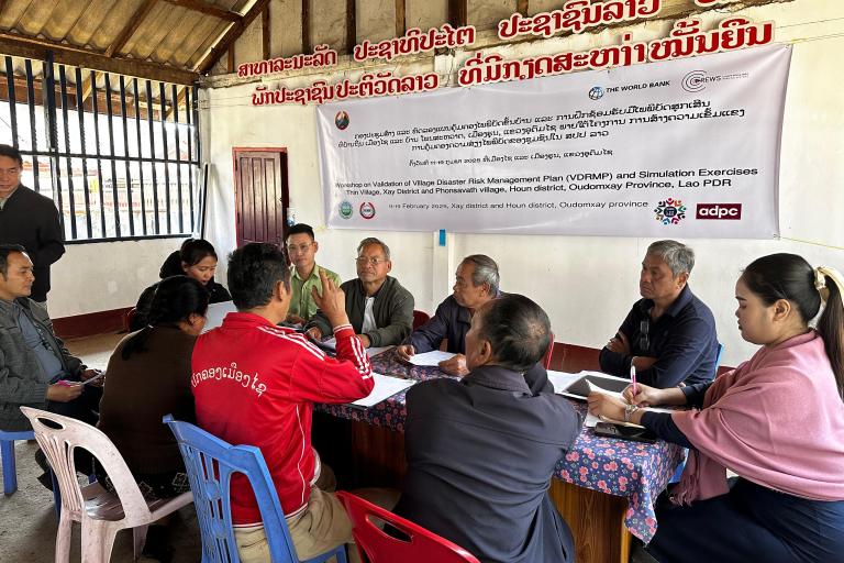 A group of people sit around tables in a meeting room, discussing documents. A banner about disaster risk management in Laos hangs on the wall behind them.