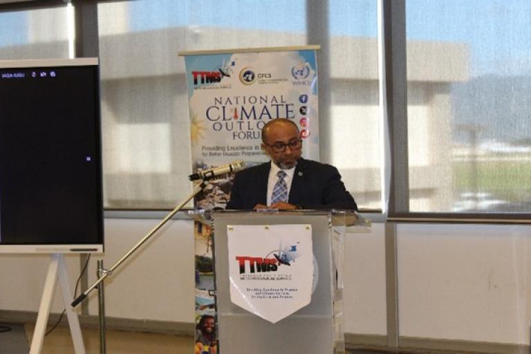 A man in a suit stands at a podium speaking during the National Climate Outlook Forum, with a presentation screen and banner in the background.